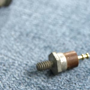May include: A close-up of a small, metallic component with a threaded end, a hexagonal body, a silver band, a brown cylindrical section, and a gold-colored tip. The component is resting on a blue surface.
