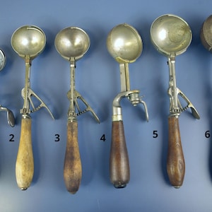 May include: A collection of six vintage ice cream scoops. Each scoop has a metal bowl and a handle, with variations in handle material and design. The scoops are arranged against a blue background, showcasing their diverse styles.