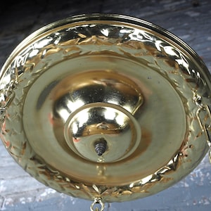 May include: A polished brass ceiling canopy with a circular shape and a decorative embossed laurel leaf border. The center features a raised dome with a small metal finial at the apex. Two gold-toned metal chains are attached to the sides.