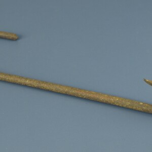 May include: A curved, rusty metal hook with a pointed end. The hook is a dark, muted gold color and lies on a blue-grey surface. The hook is likely used for hanging items.