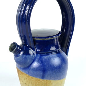Op de afbeelding: Een blauwe en bruine theepot van keramiek met een unieke handgreep die om de bovenkant van de pot wikkelt. De handgreep is gemaakt van één stuk keramiek en heeft een gladde, afgeronde vorm.