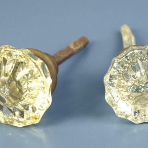 Puede incluir: Dos pomos de puerta antiguos de cristal con diseños facetados en forma de flor. Los pomos son transparentes con un ligero tinte amarillo, y los vástagos son de color marrón óxido. El fondo es azul liso y apagado.