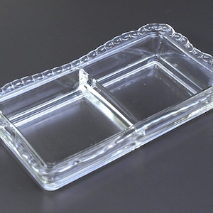 Puede incluir: Un plato de vidrio transparente rectangular dividido en dos compartimentos con borde festoneado. Ideal para servir aperitivos o condimentos. El vidrio transparente permite ver el contenido.