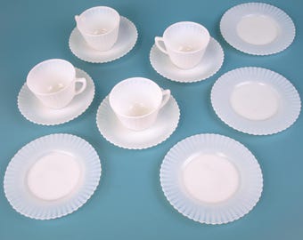 May include: A set of white teacups and saucers with a ribbed design and scalloped edges. The teacups have handles and are placed on matching saucers. Several additional saucers are also included, all arranged on a light blue surface.