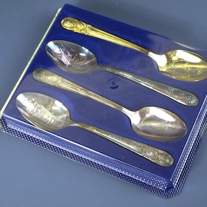 May include: A boxed set of four decorative spoons. Three are silver-toned, one is gold. Each spoon features detailed designs on the handle and bowl. The spoons are presented in a blue box with a clear plastic lid, ideal for display or gifting.