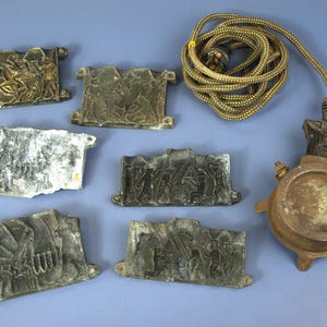 May include: A collection of six rectangular metal plates featuring embossed figural scenes, accompanied by a circular metal heating base with a coiled brown fabric-covered electrical cord and a plug, all arranged on a flat surface.