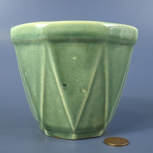 Puede incluir: Una maceta de cerámica de color verde claro con un acabado brillante y una textura de esmalte agrietado. El recipiente presenta un diseño exterior geométrico y facetado con crestas verticales. Una moneda de cobre está al lado para escala.