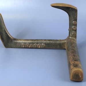 May include: An antique, cast iron cobbler's last, with the word "DRAGON" and "BON" embossed on the surface. The tool has a curved, hook-like shape and a vertical post. The metal has a dark, aged appearance.
