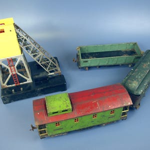 Può includere: Una collezione di vagoni di treni giocattolo vintage e una gru. La gru è gialla e argentata, con una base nera. I vagoni sono verdi e rossi, con segni di usura. Lo sfondo è blu.