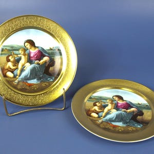 May include: Two decorative plates with gold rims and detailed artwork. The plates feature a central image of a woman and two children in a classical style. One plate is displayed on a metal stand, while the other rests flat.