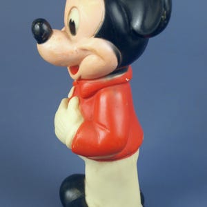May include: Vintage Mickey Mouse figurine. The figure features a black head and ears, a red jacket, white gloves, and black shoes. The figurine is standing and has a smiling expression. A classic Disney collectible.