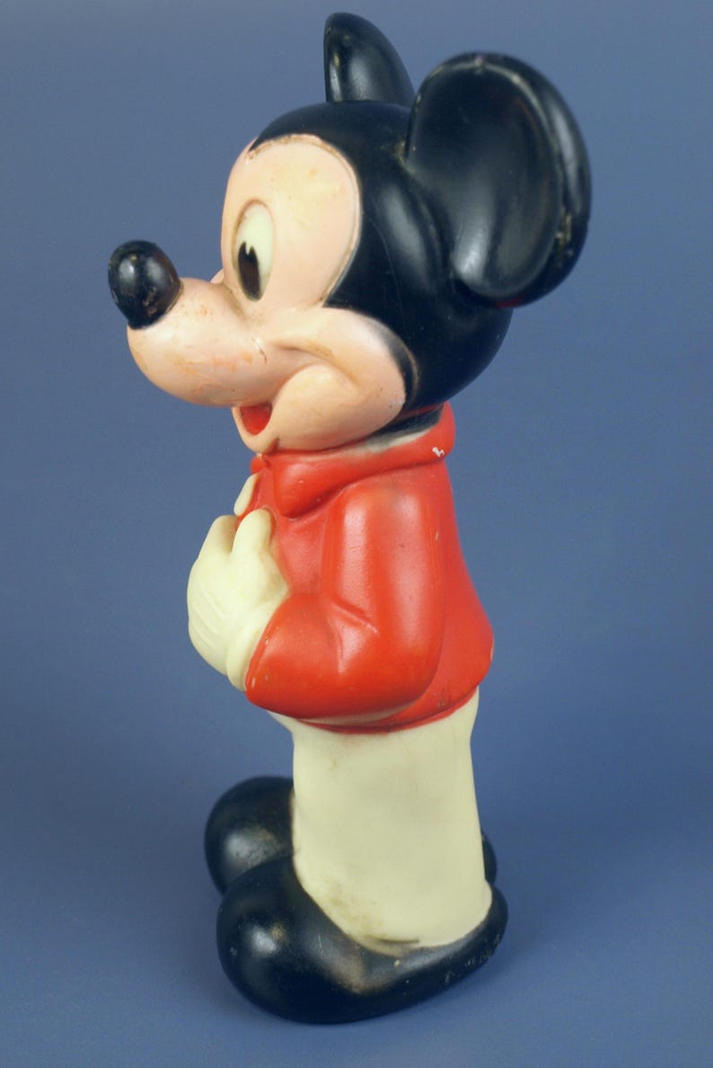 May include: Vintage Mickey Mouse figurine. The figure features a black head and ears, a red jacket, white gloves, and black shoes. The figurine is standing and has a smiling expression. A classic Disney collectible.