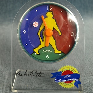 May include: A clear acrylic baseball-themed clock featuring a colorful dial with a yellow baseball player silhouette. The clock face is divided into blue and red sections, with an orange bat and hands. The base has Babe Ruth's signature and a 100th Anniversary emblem.