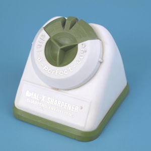 May include: White and green IAL-X sharpener with the text "a perfect edge" and "SHARPENS EVERYTHING". The sharpener has a white body with a green top and base. The background is a solid blue.
