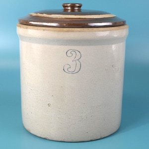 Pode incluir: Um pote de grés cilíndrico com corpo bege claro e tampa esmaltada em castanho escuro. A frente do pote apresenta um número 3 azul pintado à mão. O recipiente tem uma borda espessa e um acabamento mate liso na superfície exterior.