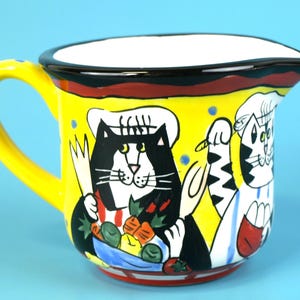 May include: A yellow ceramic pitcher with a black rim and a yellow handle. The pitcher is decorated with cartoon cats in chef hats, holding utensils and vegetables. The background is a solid light blue.