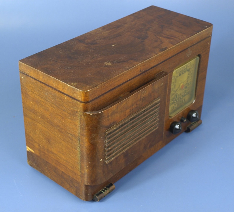 Puede incluir: Una radio vintage de madera con un gabinete de madera marr&oacute;n y una rejilla de altavoz negra. La radio tiene un dial con una cara de vidrio y algunas perillas en la parte delantera.