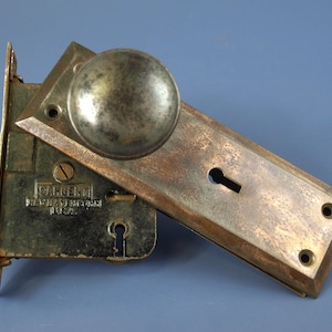 Puede incluir: Un pomo de puerta de metal vintage con placa rectangular y acabado desgastado. El mecanismo de cerradura interno es visible, con el texto SARGENT NEW HAVEN CONN U.S.A. y una cerradura. Los herrajes presentan una pátina envejecida.