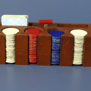 May include: A rectangular wooden poker chip rack with four vertical slots containing stacks of white, red, and blue circular chips. A deck of playing cards sits in a slot on the left, and a small red handle is positioned on the top center of the rack.