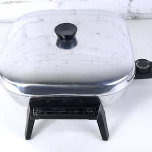 Puede incluir: Una sartén eléctrica cuadrada con acabado de metal plateado pulido y tapa a juego. La tapa cuenta con un pomo circular negro. La base tiene patas de plástico negro y un dial de control de temperatura negro con cable de alimentación.