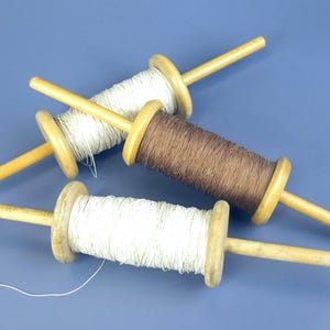 May include: Three wooden kite spools with string, arranged on a blue surface. Two spools are wound with white string, and one is wound with brown string. The spools have a light wood color.