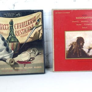 May include: Two vintage vinyl record albums. One album cover is black with a wine bottle, a hat, and text in a banner. The other album cover is red with a painting and the title "Rigoletto".