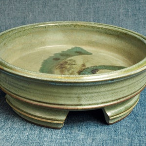 May include: A green ceramic planter with a wide, round base and three legs. The planter has a glazed finish with a subtle green and brown pattern.