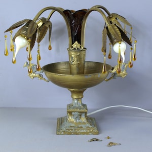 May include: A gold-toned metal lamp with a decorative base and a central bowl. The lamp has multiple arms with leaf-shaped accents and dangling amber glass beads. The lamp has a dark amber glass shade and features horse-shaped accents on the base.