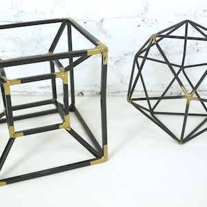 May include: Two geometric sculptures made of thin black metal rods with gold-colored corner connectors. One sculpture is a nested cube structure, while the other is a multi-faceted icosahedron shape. Both objects feature a wireframe design.