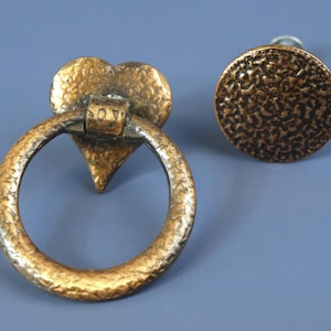 Puede incluir: Dos herrajes de gabinete antiguos de bronce. Uno es una placa trasera en forma de corazón con un tirador de anillo, el otro es un pomo redondo. Ambos tienen una textura martillada y un acabado cálido y envejecido. El tirador de anillo tiene la palabra "LOVE".