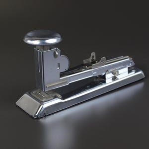 May include: A shiny, chrome-plated stapler with a rounded top and a rectangular base. The stapler is designed for office or school use and is shown against a dark background.
