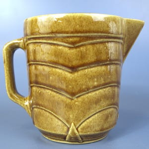 May include: A ceramic pitcher with a glossy mustard-brown glaze and a textured surface featuring repeating horizontal chevron patterns. The vessel includes a sturdy side handle and a pointed pouring spout, showcasing a uniform color finish.