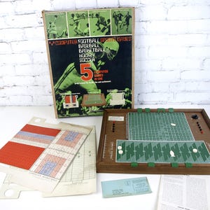 Może przedstawiać: Zabytkowa gra planszowa "Computer Football Sports Games". Pudełko zawiera ilustracje futbolu amerykańskiego, baseballu, koszykówki, hokeja i piłki nożnej. Gra zawiera drewnianą planszę, arkusze instrukcji i elementy gry.