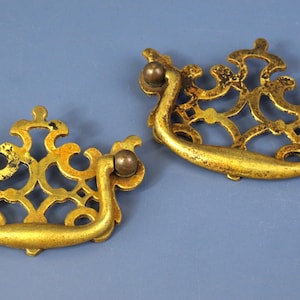 May include: Two antique brass drawer pulls with ornate, openwork designs. Each pull has a curved handle and decorative end pieces. The brass has a warm, aged patina, and the pulls are set against a blue background.