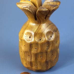 May include: A carved wooden pineapple sculpture with a textured surface and a golden-brown finish. The pineapple has a detailed crown and a series of small, evenly spaced holes. A penny is next to the pineapple for scale.