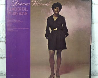 Op de afbeelding: Vintage vinyl albumhoes met Dionne Warwick. De albumhoes heeft een paarse verticale streep aan de linkerkant met de tekst "I'll Never Fall in Love Again". Dionne Warwick is afgebeeld in een zwarte jurk met een gouden riem.