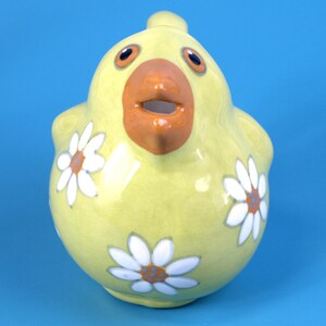 Puede incluir: Una jarra de cer&aacute;mica amarilla en forma de pollo, con una gran boca naranja y ojos grandes. La jarra est&aacute; decorada con dise&ntilde;os de flores de margarita blancas con centros naranjas y azules. El fondo es azul liso.