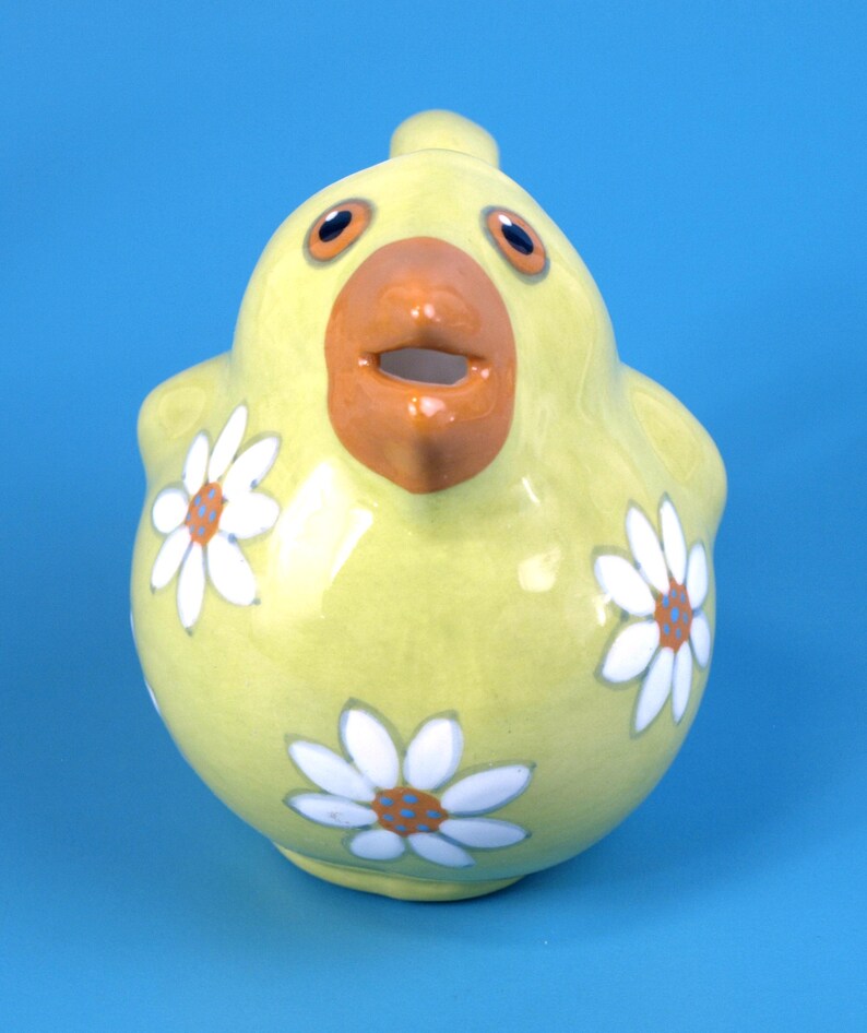 Puede incluir: Una jarra de cer&aacute;mica amarilla en forma de pollo, con una gran boca naranja y ojos grandes. La jarra est&aacute; decorada con dise&ntilde;os de flores de margarita blancas con centros naranjas y azules. El fondo es azul liso.