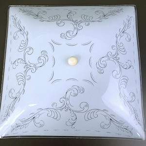 Puede incluir: Luminaria de techo cuadrada de vidrio esmerilado con un diseño decorativo de volutas negras. La luz tiene un accesorio central redondo de color crema y un borde de metal plateado. La luz es de estilo vintage.