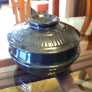 Puede incluir: Un cuenco negro de cerámica con tapa y acabado brillante. La tapa presenta un pequeño asa y detalles acanalados verticales. El cuenco está sobre una superficie de vidrio, reflejando la luz y los objetos circundantes.