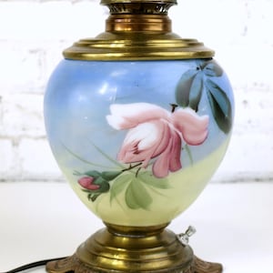 Może przedstawiać: Antyczna lampa naftowa z ręcznie malowanym kloszem ze szkła przedstawiającym różową różę i zielone liście na niebieskim i żółtym tle. Lampa ma mosiężną podstawę i mosiężny komin.
