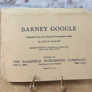 May include: A vintage book cover for a Barney Google comic book. The cover is brown with black text that reads "Barney Google", "Adapted from the famous Newspaper Strip", "By BILLY DeBECK", "Copyright MCMXXXV, King Features Syndicate, Inc. Great Britain rights reserved", "Published by THE SAALFIELD PUBLISHING COMPANY", "AKRON, OHIO", "Publishers for the Children", "Made in U.S.A.", and "NEW YORK".