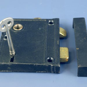 May include: A vintage black metal lock with a silver key inserted. The lock has brass-colored bolts and is accompanied by a matching black metal strike plate. The lock and strike plate are set against a blue background.