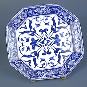 Könnte beinhalten: Eine achteckige Keramikplatte mit weißem Grund und aufwendigen blauen Blumenmustern. Der Rand der Platte weist ein zartes Blumendesign auf, und die Mitte zeigt ein geometrisches und florales Motiv. Die Platte wird auf einem kleinen Ständer präsentiert.