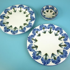 Puede incluir: Dos platos redondos de cerámica y un cuenco pequeño de cerámica, todos con una base de color crema y un patrón pintado a mano de flores azules y tallos verdes dispuestos en un borde circular alrededor de cada pieza.