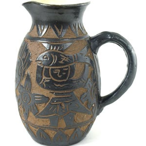 May include: A handmade ceramic pitcher with a black glaze and brown carved designs. The pitcher features a handle and a spout. The carved designs include fish, floral patterns, and geometric shapes. The interior of the pitcher is a light cream color.