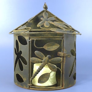 May include: A cylindrical metal lantern with a dark, weathered bronze finish featuring cut-out dragonfly and leaf motifs. The structure includes a hinged door with a latch and a peaked roof. A cream-colored pillar candle sits inside the lantern.