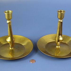 May include: Two brass candle holders with round bases and a single candle holder on each. The candle holders are on a blue surface.