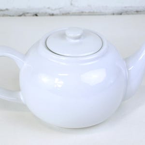 Puede incluir: Una tetera de cerámica blanca con cuerpo redondeado, asa curva y boquilla. La tapa tiene un pequeño pomo. La tetera está diseñada para preparar té y tiene un acabado brillante. Es un artículo de menaje.