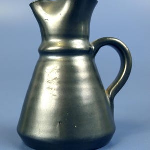May include: A small, dark gray ceramic pitcher with a handle and a spout. The pitcher has a rounded base that tapers to a wider top. The handle is a simple loop. The surface has a slightly textured appearance.